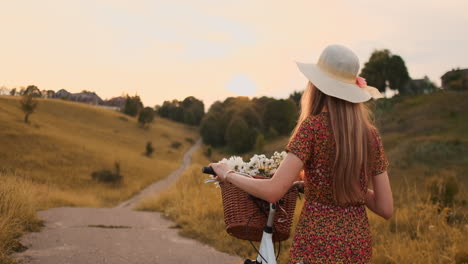 Back-plan-slow-motion:-a-Beautiful-blonde-in-a-dress-with-flowers-in-a-basket-and-a-retro-bike-walks-along-the-road-in-the-summer-field-looking-around-and-smiling-feeling-free.
